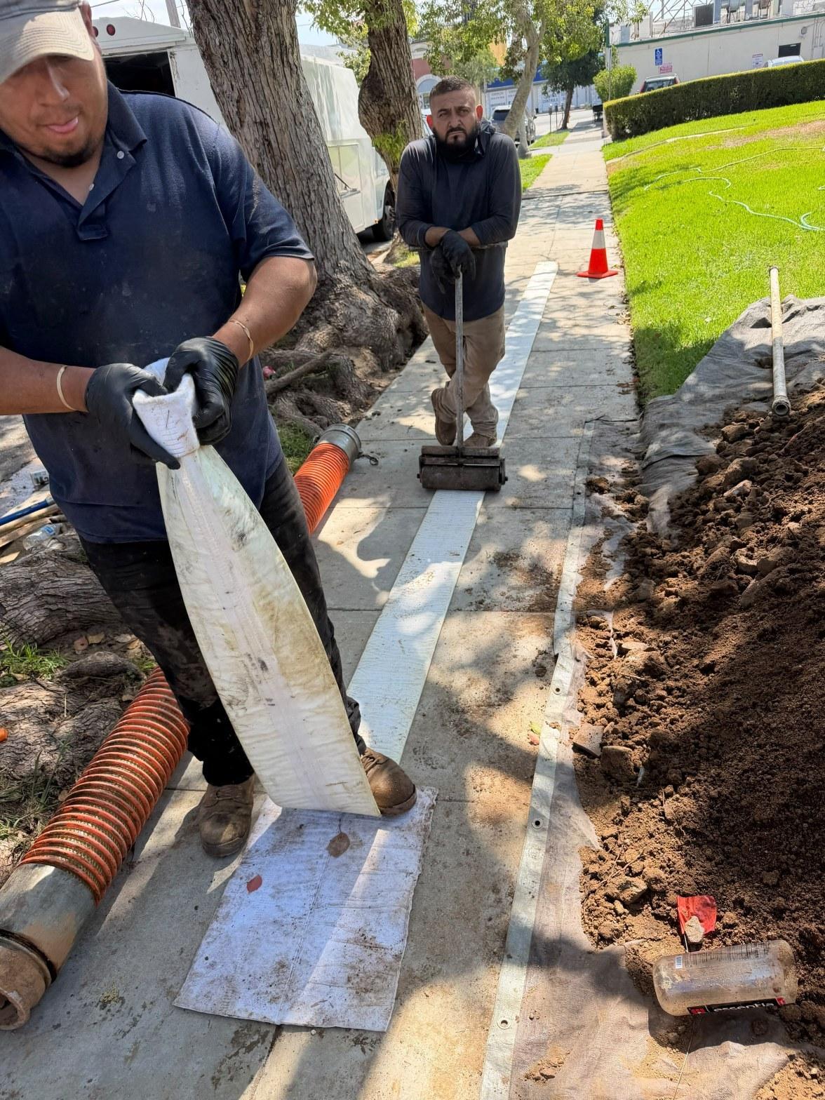 Workers installing underground pipe in sidewalk trench with vacuum hose