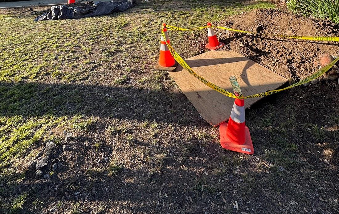 Excavated sewer trench covered with plywood and surrounded by orange safety cones and yellow caution tape in a residential front yard